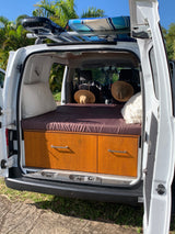 This RV rental campervan shows its comfortable bed, built-in wooden storage, and a surfboard on the roof rack, ready for trips.