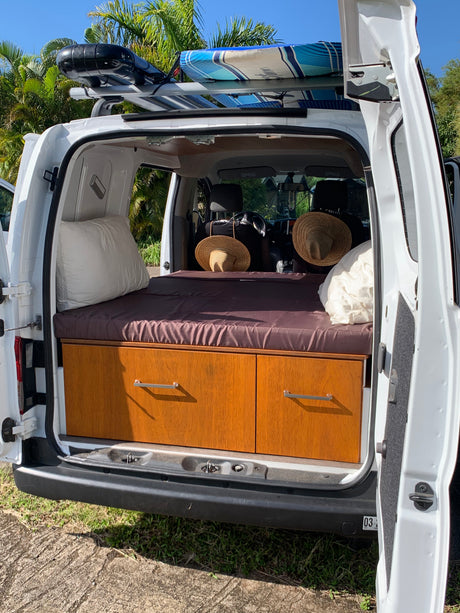 This RV rental campervan shows its comfortable bed, built-in wooden storage, and a surfboard on the roof rack, ready for trips.