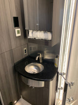A compact RV rental bathroom with a silver sink, dark wood cabinets, a shelf of white towels, and a digital thermostat.
