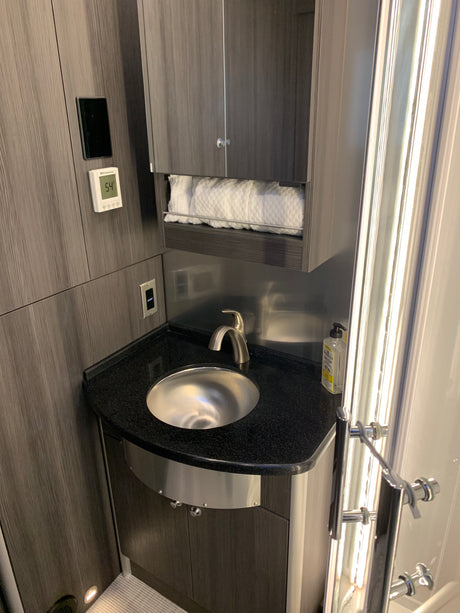 A compact RV rental bathroom with a silver sink, dark wood cabinets, a shelf of white towels, and a digital thermostat.
