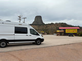 White RV rental van parked at Devils Tower KOA Cafe & Gifts with Devils Tower in the background under an overcast sky.