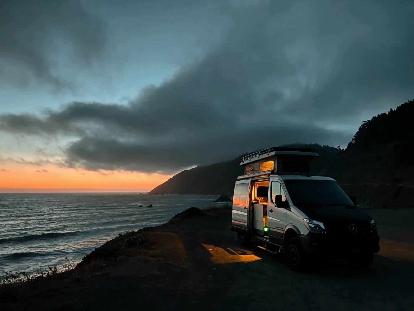 An RV rental camper van at a dramatic coastal sunset, warm light from inside illuminates the ocean overlook.