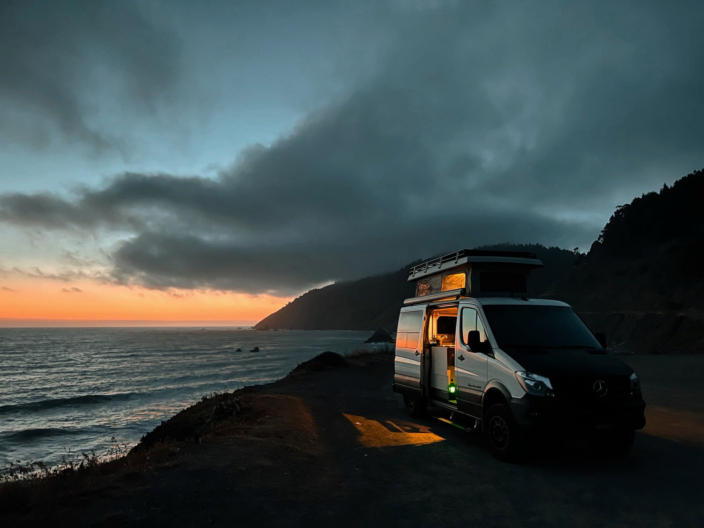 An RV rental camper van at a dramatic coastal sunset, warm light from inside illuminates the ocean overlook.