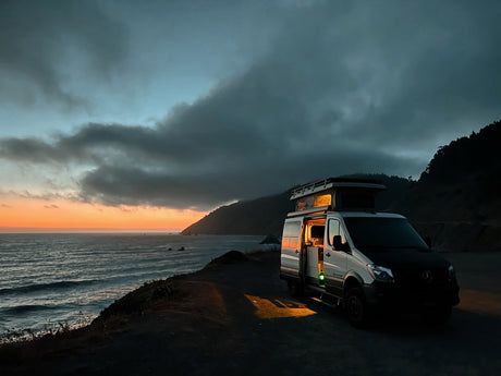 An RV rental camper van at a dramatic coastal sunset, warm light from inside illuminates the ocean overlook.