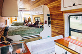 Cozy RV rental interior with a woman reading in bed, a baby crib, and a small kitchen area. Ideal for mobile living.