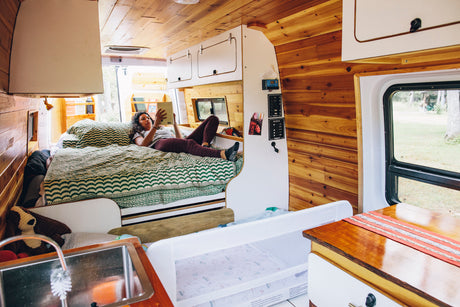 Cozy RV rental interior with a woman reading in bed, a baby crib, and a small kitchen area. Ideal for mobile living.