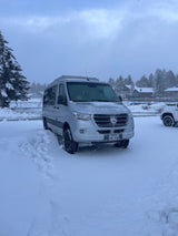 A silver Mercedes-Benz Sprinter RV rental is parked in deep snow on a cloudy winter day.