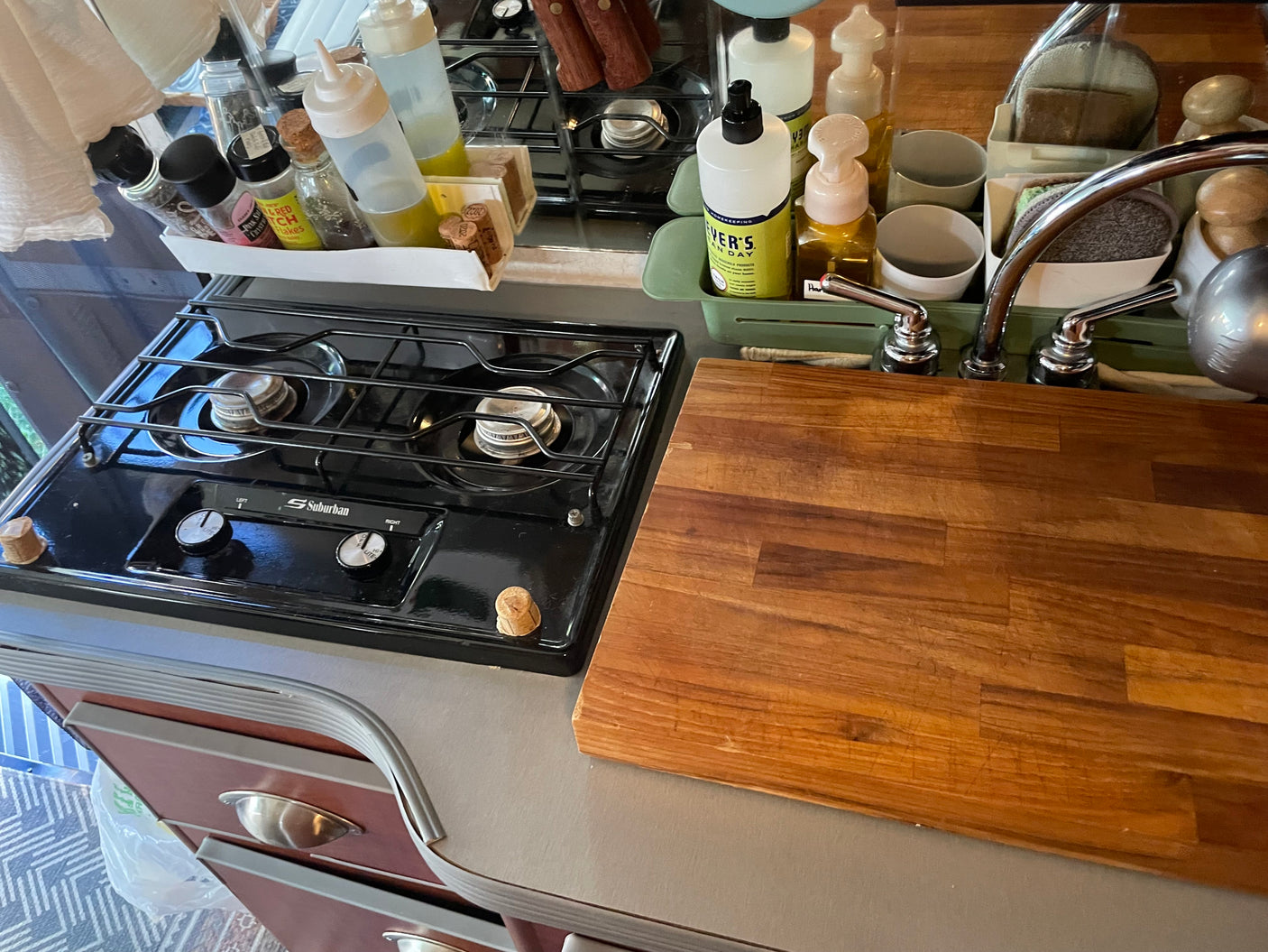 Compact RV rental kitchen features a Suburban stove, sink, and prep area with a wooden cutting board & cooking supplies.