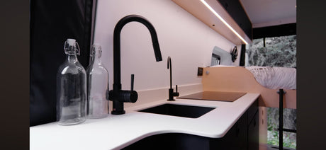 Sleek RV rental kitchen area: black sink, faucet, induction cooktop, white counters, and glass bottles. Compact and functional.
