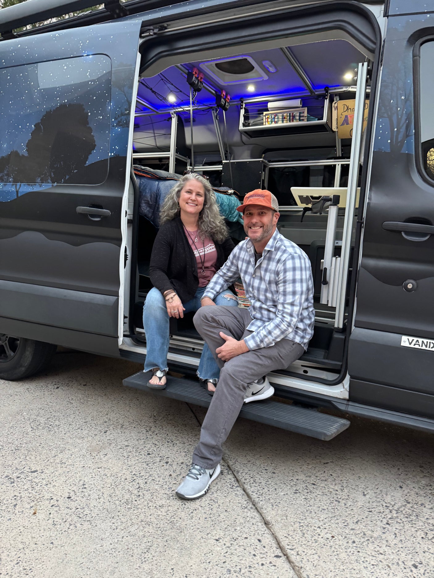 Smiling couple sits inside their customized camper van RV rental, showcasing its unique starry exterior and cozy blue-lit interior.