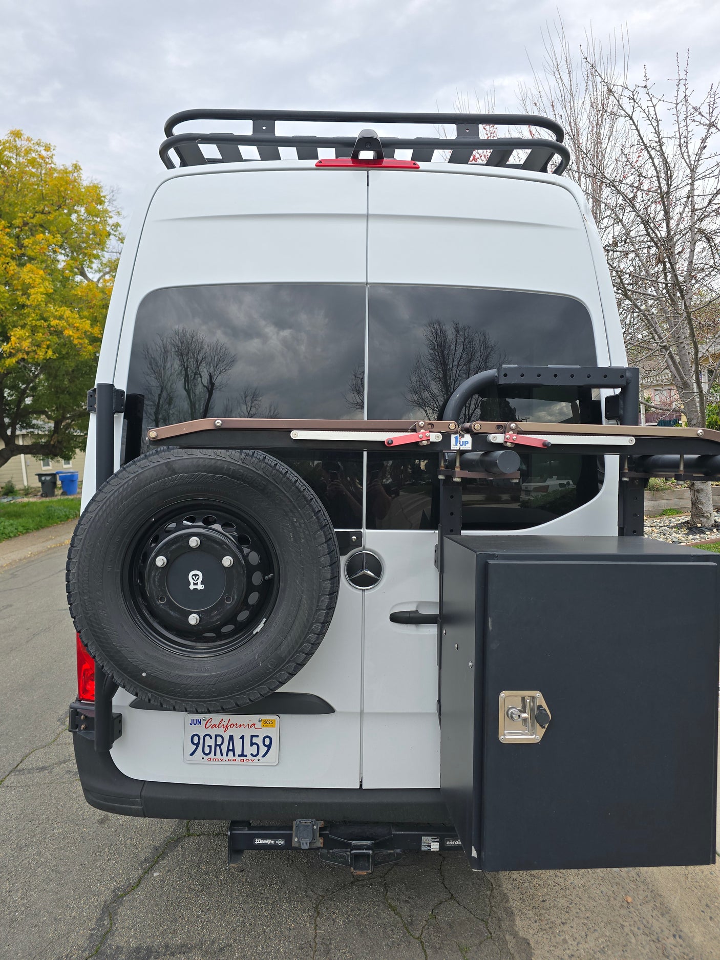 Explore with this white Sprinter RV rental van, featuring a roof rack, rear spare tire, storage box, and bike rack.
