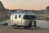 A silver Airstream travel trailer RV rental rests by a calm lake, surrounded by desert mountains under a soft twilight sky.