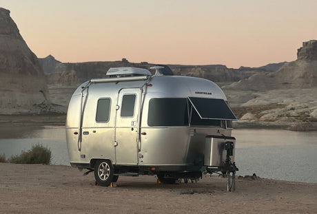 A silver Airstream travel trailer RV rental rests by a calm lake, surrounded by desert mountains under a soft twilight sky.