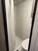 View of a compact RV rental bathroom featuring a white shower curtain, shower pan, and toilet against grey tiled walls.