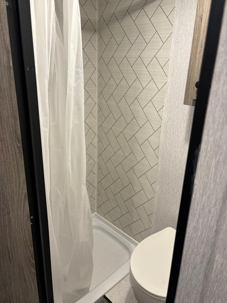 View of a compact RV rental bathroom featuring a white shower curtain, shower pan, and toilet against grey tiled walls.