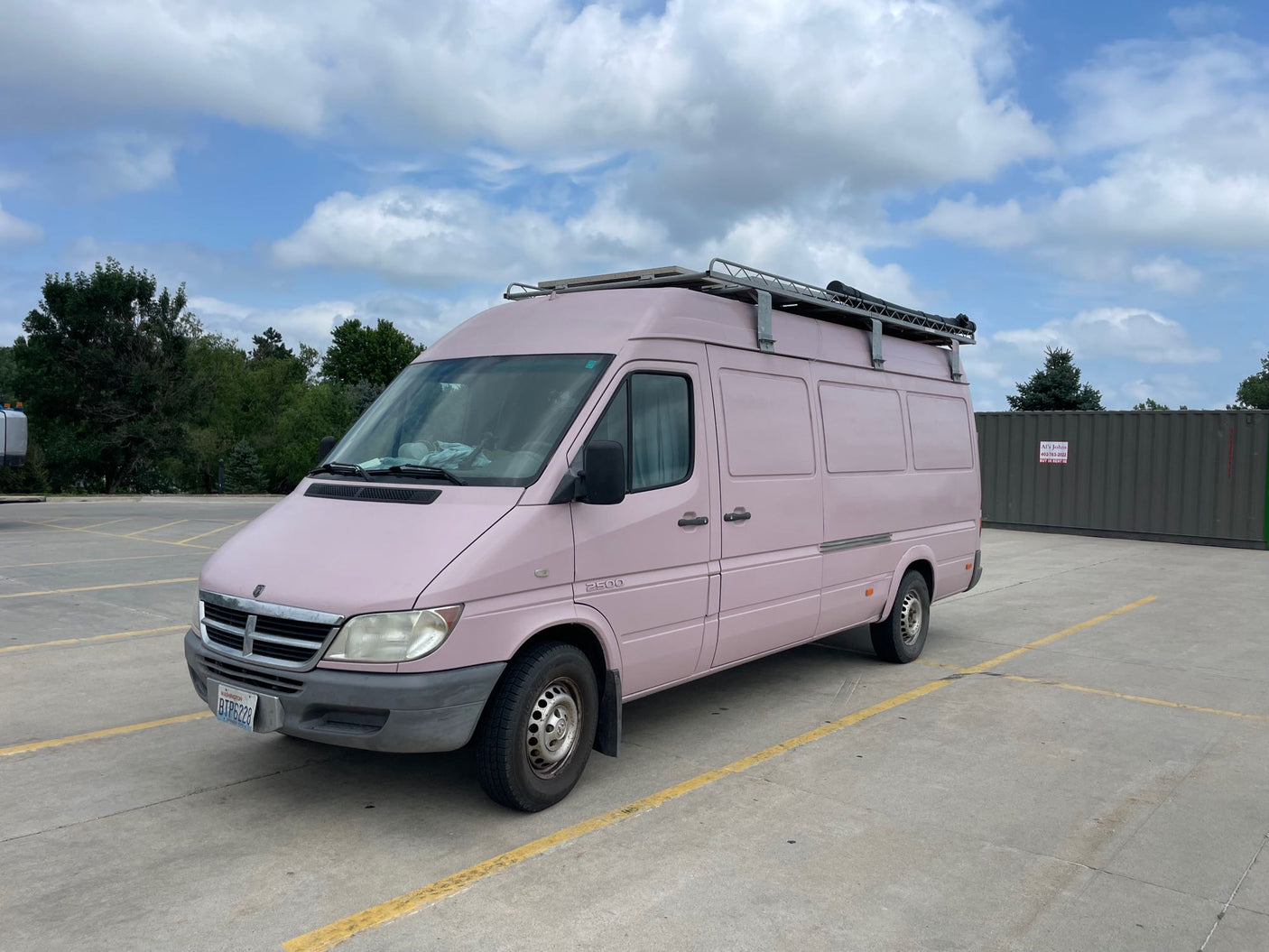 A light pink Dodge Sprinter camper van with a roof rack, likely a custom RV rental conversion, parked in a lot.