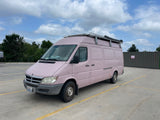 A light pink Dodge Sprinter camper van with a roof rack, likely a custom RV rental conversion, parked in a lot.
