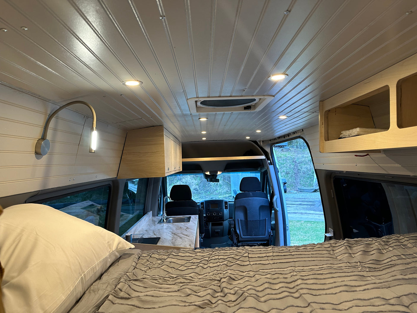 Bright RV rental interior: A cozy bed in the foreground, facing a kitchenette, storage, and cockpit. Beaded ceiling with lights.