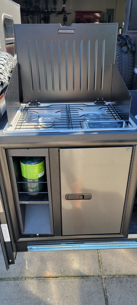 RV rental's slide-out outdoor kitchen with an open Dometic 2-burner gas stove, visible propane tank, and mini-fridge.
