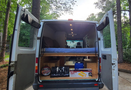 View into an RV rental camper van's open rear doors, revealing a raised bed, tools, jumper cables, and RV levelers in storage.