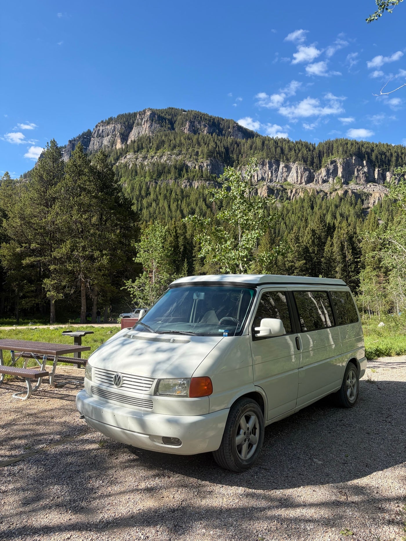 White camper van RV rental at a serene mountain campsite, surrounded by pine forests and rocky cliffs under blue skies. — photo 4