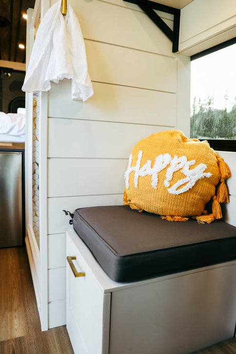Cozy rv rental interior: white shiplap walls, a comfy dark gray bench cushion, and a cheerful "Happy" pillow.