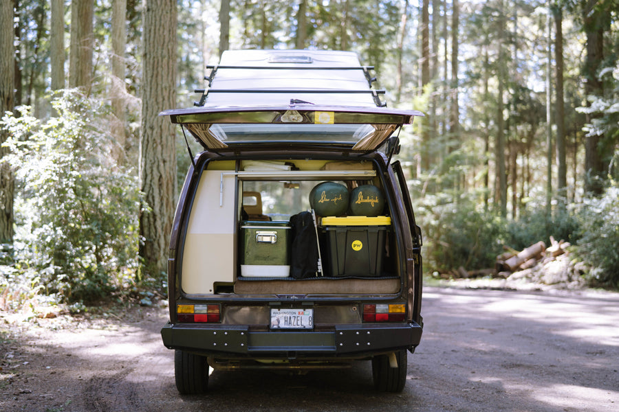 A brown camper van named 'Hazel' (WA plate) with its rear hatch open, loaded with camping gear, parked in a forest. Perfect for an RV rental adventure.