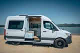 A white rv rental camper van, door open to show its sleek interior, parked on a sandy shore by a calm lake with hills.