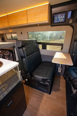 This RV rental's camper van interior features black bench seating, an adjustable table, overhead wood cabinets, and a digital control panel.
