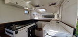 Clean and modern RV rental interior featuring a compact kitchen with sink and fridge, overhead cabinets, and a sleeping area.