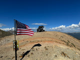 Off-road adventure with an RV rental: a Jeep with a rooftop tent on a rocky mountain peak, American flag in foreground.