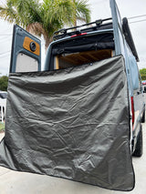 This RV rental camper van features open rear doors with a grey privacy screen, interior speaker, wood details, and roof rack lights.
