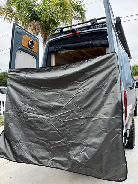 This RV rental camper van features open rear doors with a grey privacy screen, interior speaker, wood details, and roof rack lights.