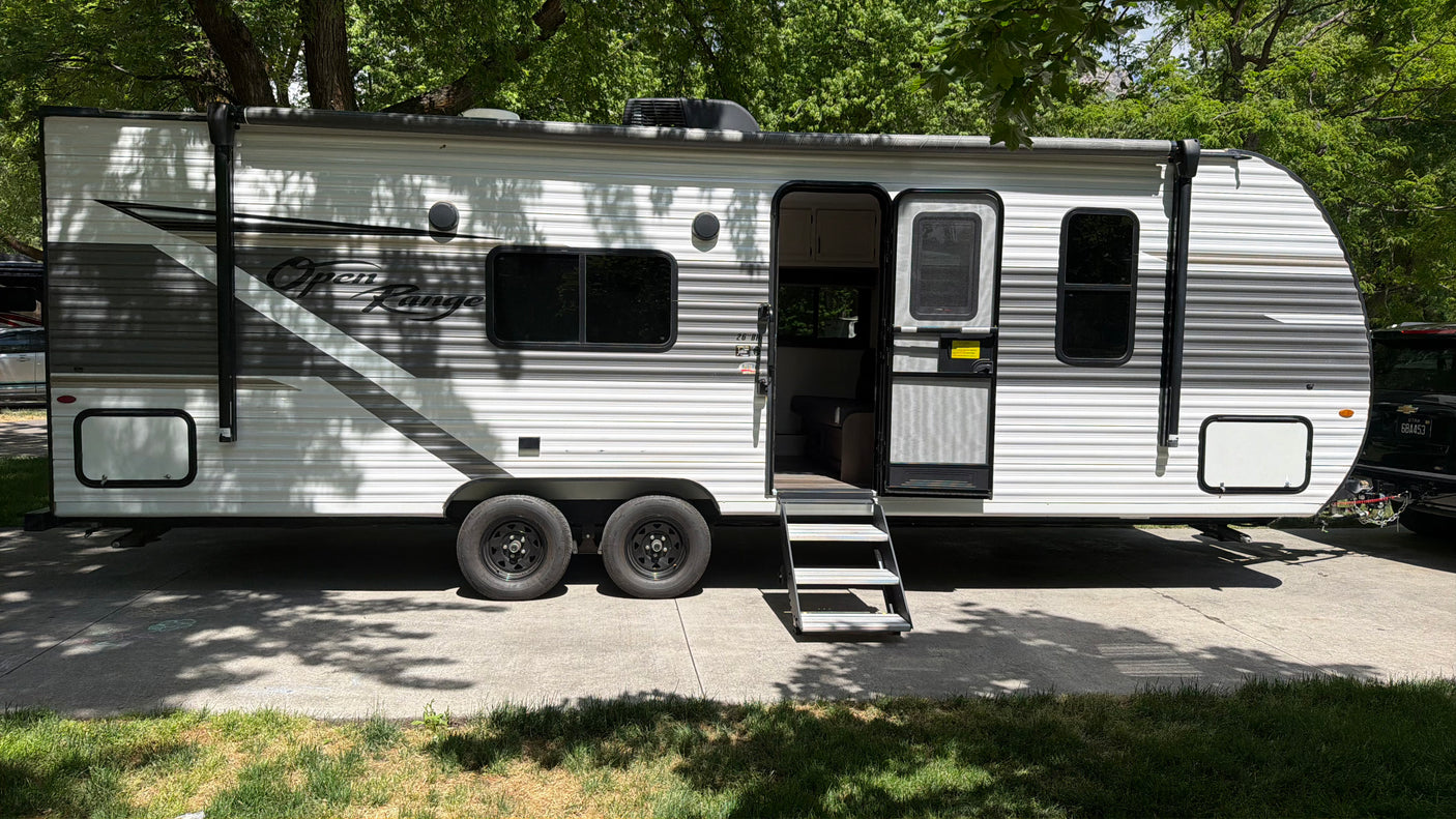 This Open Range RV rental, a white travel trailer, is parked under lush green trees, ready for your next camping trip.