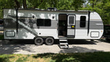 This Open Range RV rental, a white travel trailer, is parked under lush green trees, ready for your next camping trip.