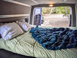 Cozy RV rental interior with bed and open rear doors revealing a natural campsite, perfect for van life adventures.
