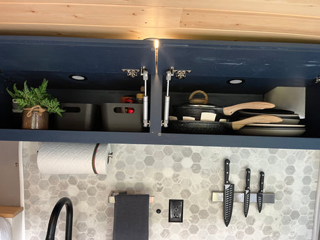 An organized RV rental kitchen displays open navy cabinets with cookware, storage, paper towels, and a hex tile backsplash.