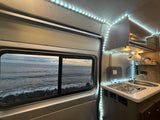 Inside an RV rental, showing the kitchen area and a scenic window view of the ocean & rocky shore.
