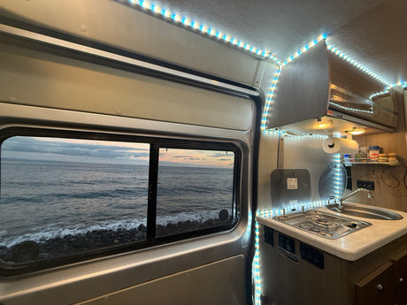 Inside an RV rental, showing the kitchen area and a scenic window view of the ocean & rocky shore.