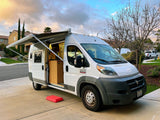 Experience van life! This white Ram ProMaster camper van, an ideal rv rental, shows its open, cozy interior and extended awning.