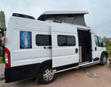 A white camper van, possibly an RV rental, with a pop-top roof and open side door. A Virgin Mary decal adorns its rear. Ideal for desert adventures.
