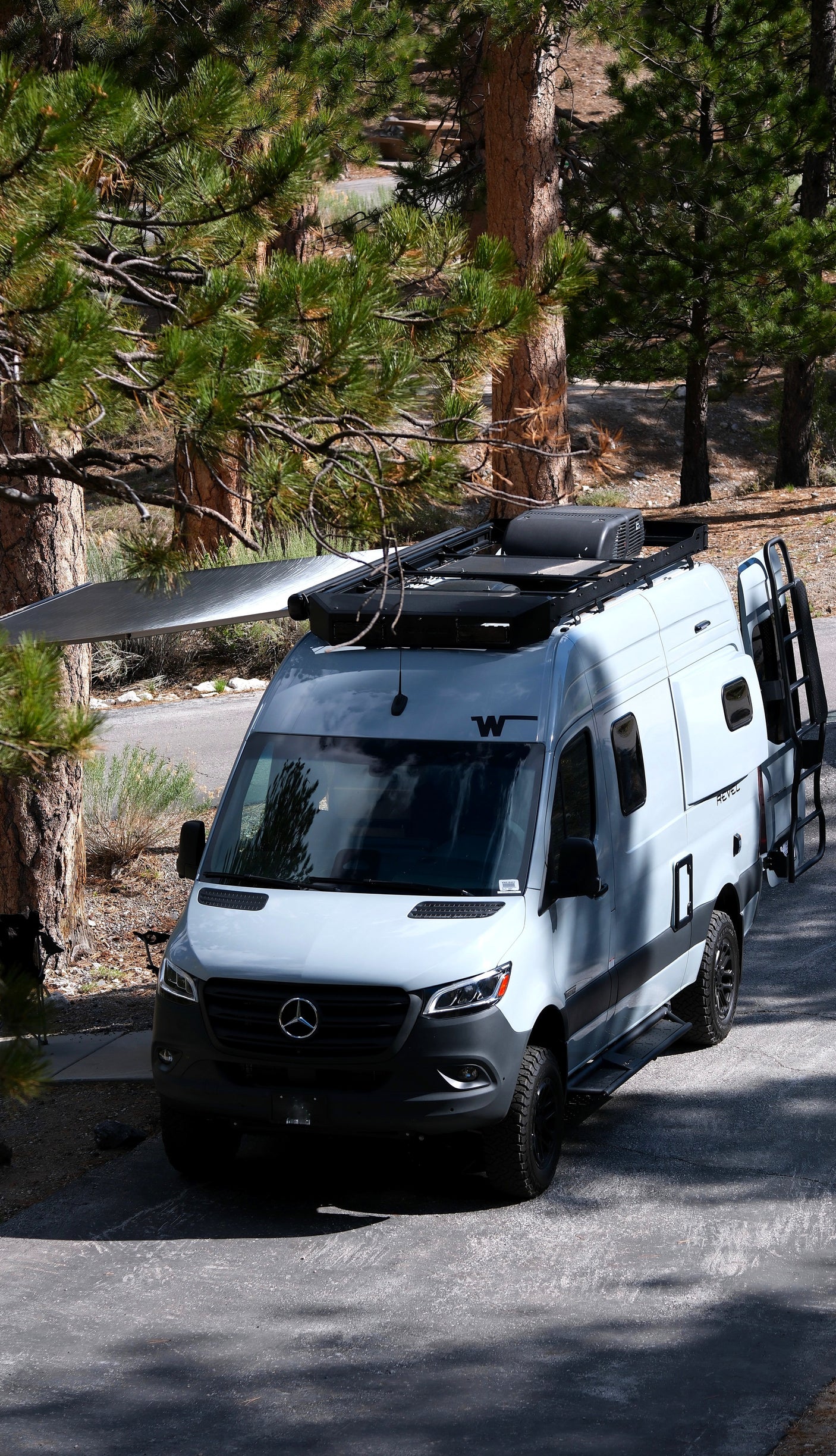 This grey RV rental, a camper van, is parked under tall pine trees with its awning extended, ready for adventure.