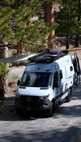 This grey RV rental, a camper van, is parked under tall pine trees with its awning extended, ready for adventure.