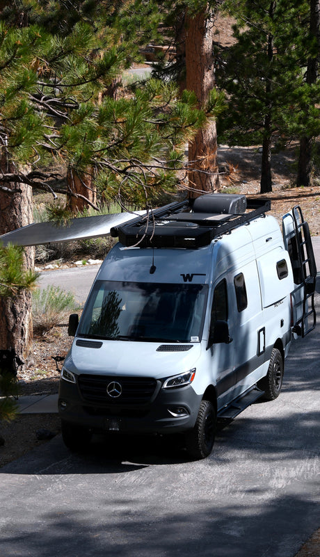 This grey RV rental, a camper van, is parked under tall pine trees with its awning extended, ready for adventure.