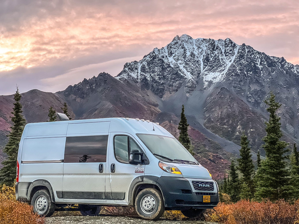 White camper van (rv rental) parked in front of snow-capped mountains and autumn foliage under a pink sky.