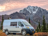 White camper van (rv rental) parked in front of snow-capped mountains and autumn foliage under a pink sky.