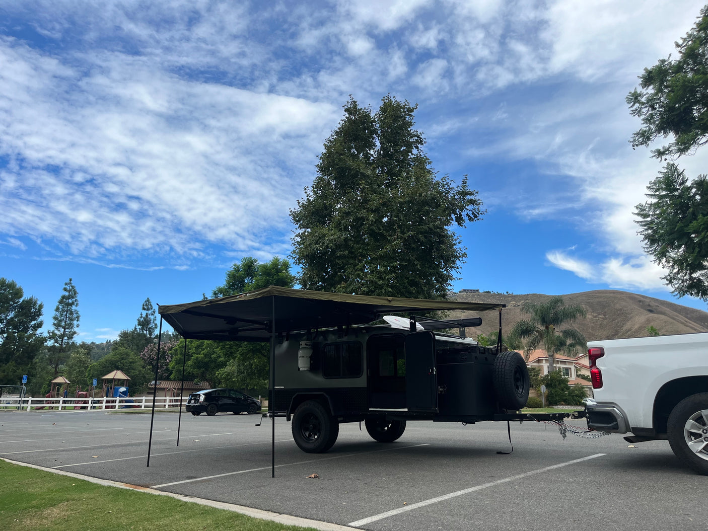 An RV rental: rugged off-road camper trailer, hitched to a white pickup in a park's lot, awning extended under a cloudy sky.
