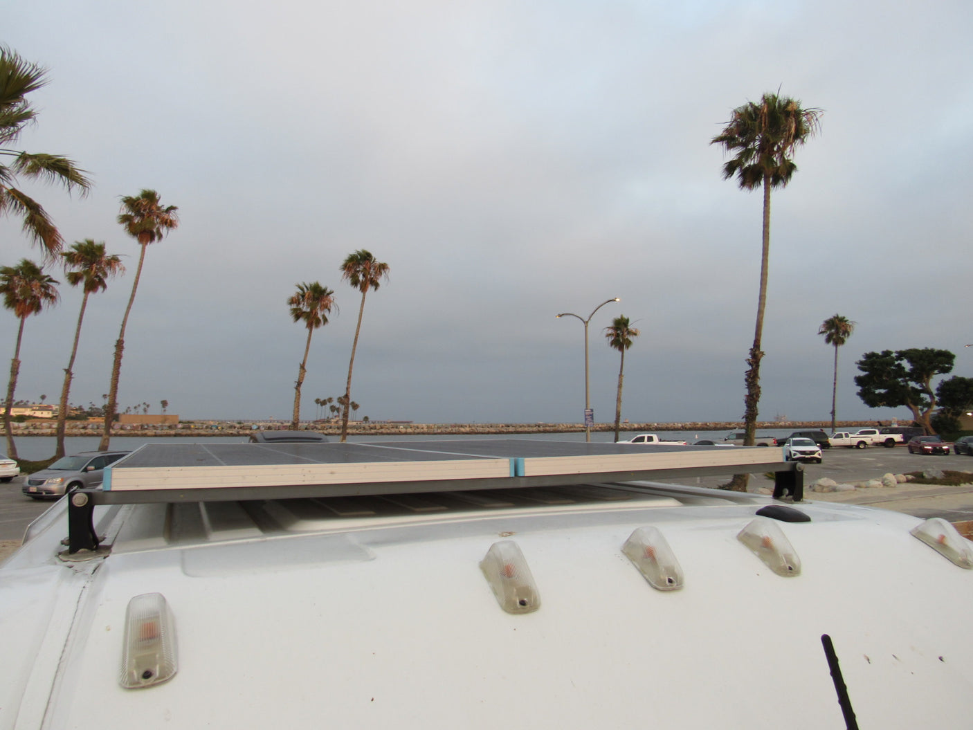 Rooftop solar panel on an RV rental, with palm trees, parked cars, and water visible under an overcast sky.