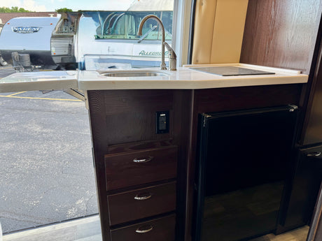 Compact RV rental kitchen with sink, stovetop, fridge, & drawers. Another travel trailer and motorhome are visible outside.