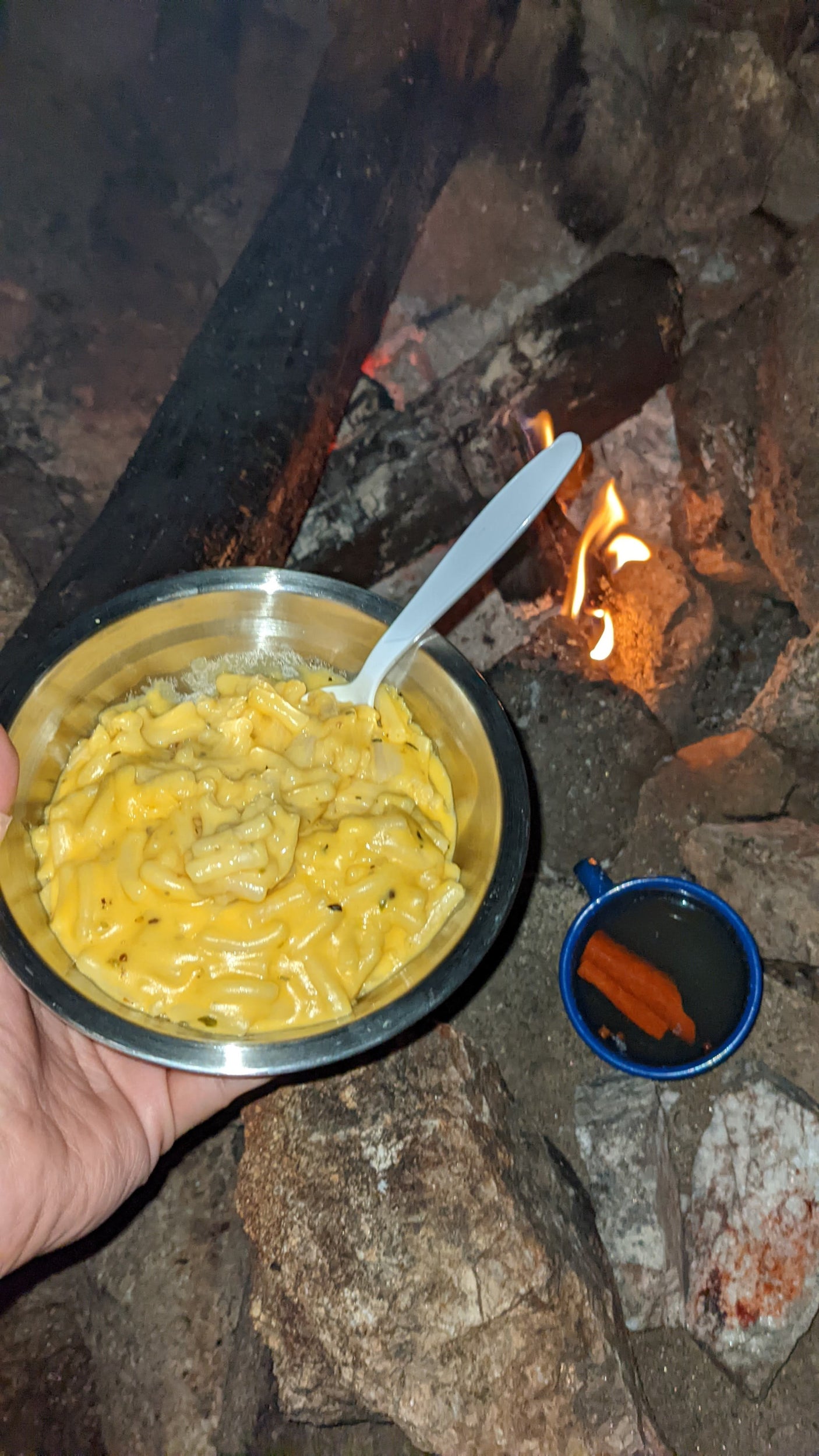 Cozy campfire scene: A person holds mac & cheese, a mug sits nearby. Perfect for an RV rental getaway!
